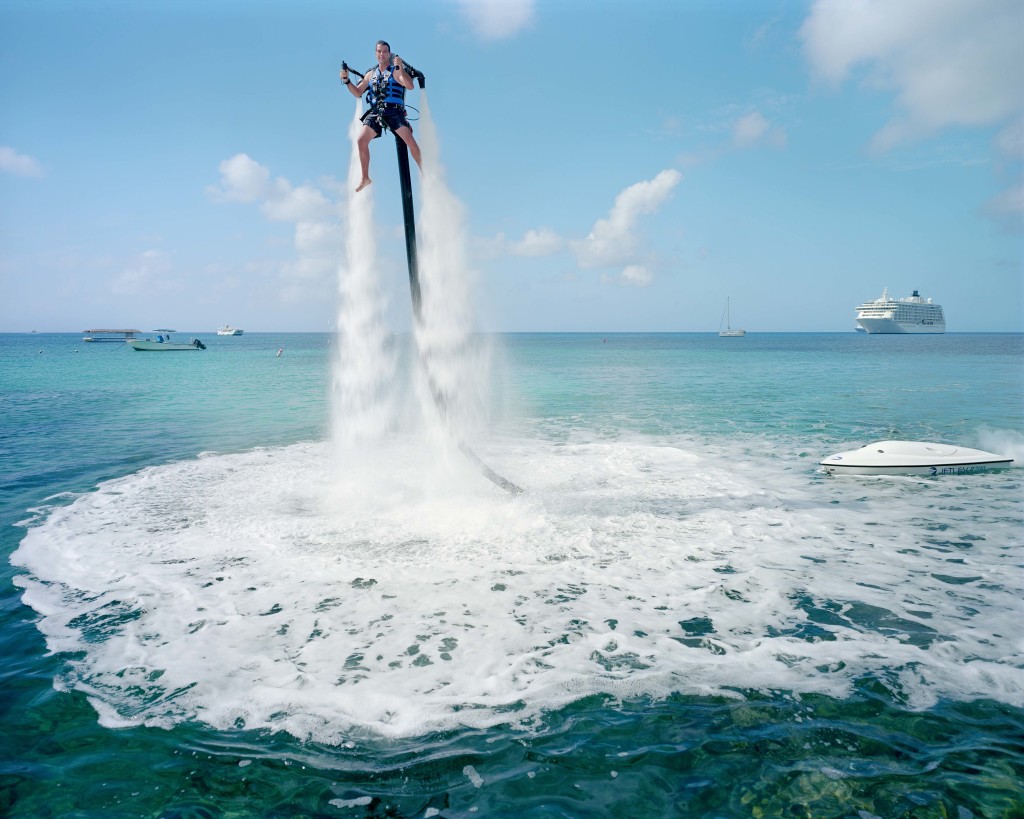An employee of “Jetpack Cayman” demonstrates this new watersport, now available on the island. A 2000cc motor pumps water up through the Jetpack, propelling the client out of the sea (359 USD for a 30-minute session). Mike Thalasinos, the owner of the company, remarks, “The Jetpack is zero gravity, the Cayman are zero taxes, we are in the right place!” Grand Cayman.
